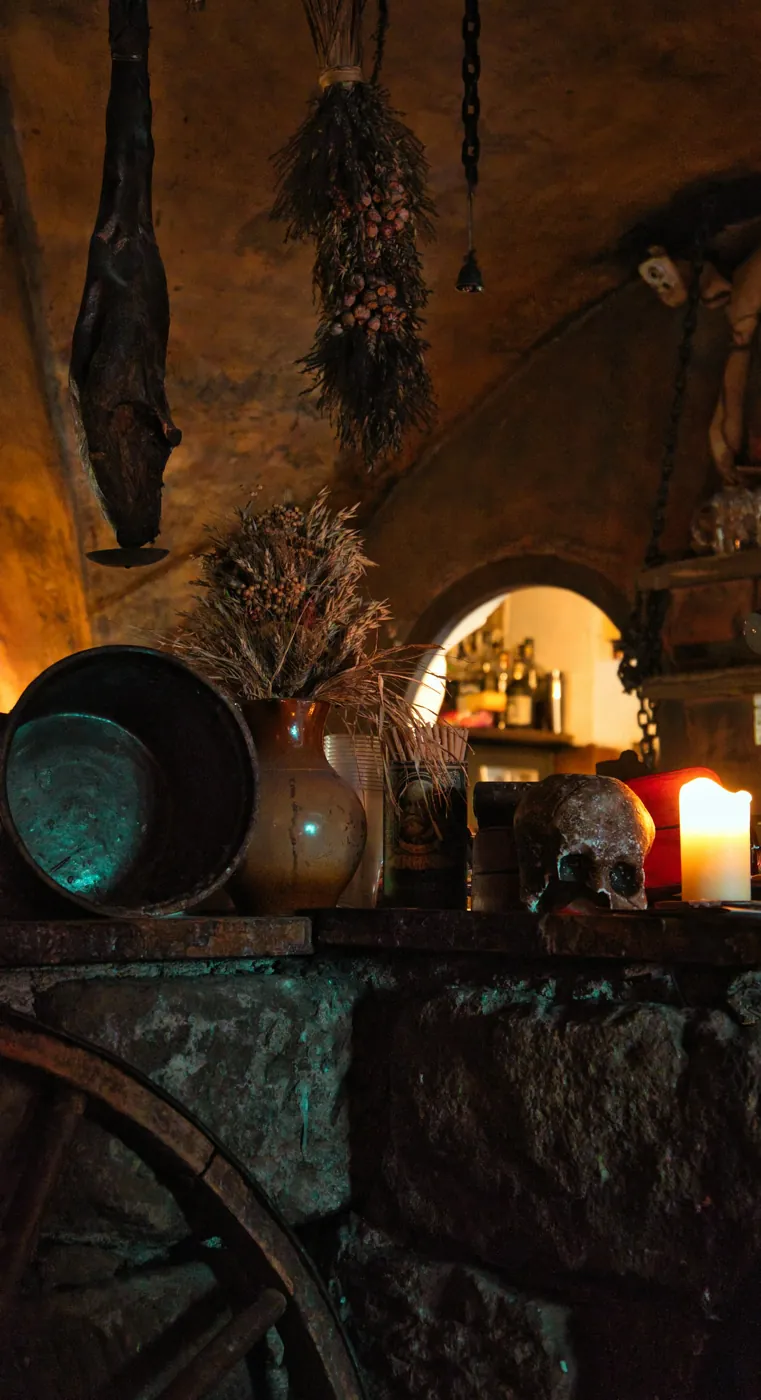 Gewölbekeller einer Prager Taverne im Kerzenlicht mit Steinmauer, Tonkrug, getrockneten Kräutern, Totenkopf-Dekoration und altem Wagenrad