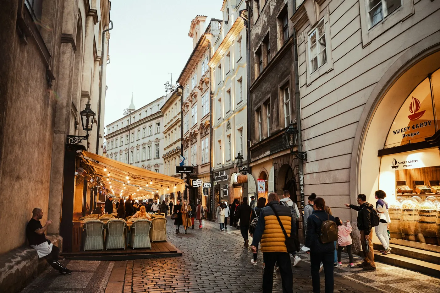 Fußgänger in einer gepflasterten Gasse der Prager Altstadt am frühen Abend, links eine Restaurantterrasse mit Lichterketten